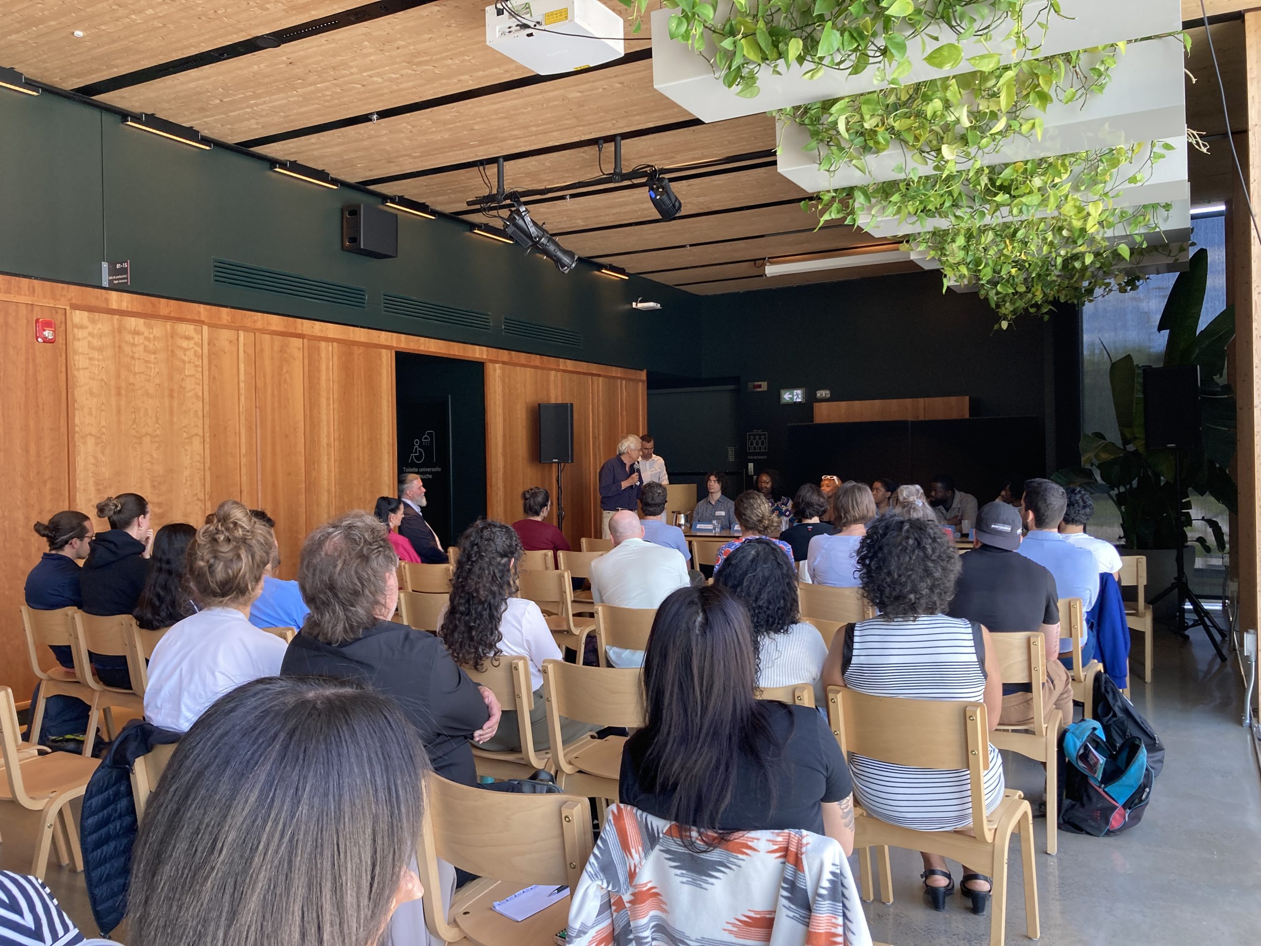 Vue sur le public de dos présent à l'événement « Que retenir de 25 ans d'intervention psychosociale par le sport? » à l'Esplanade tranquille. La moitié du plafond est couverte de plantes et les murs sont en bois.