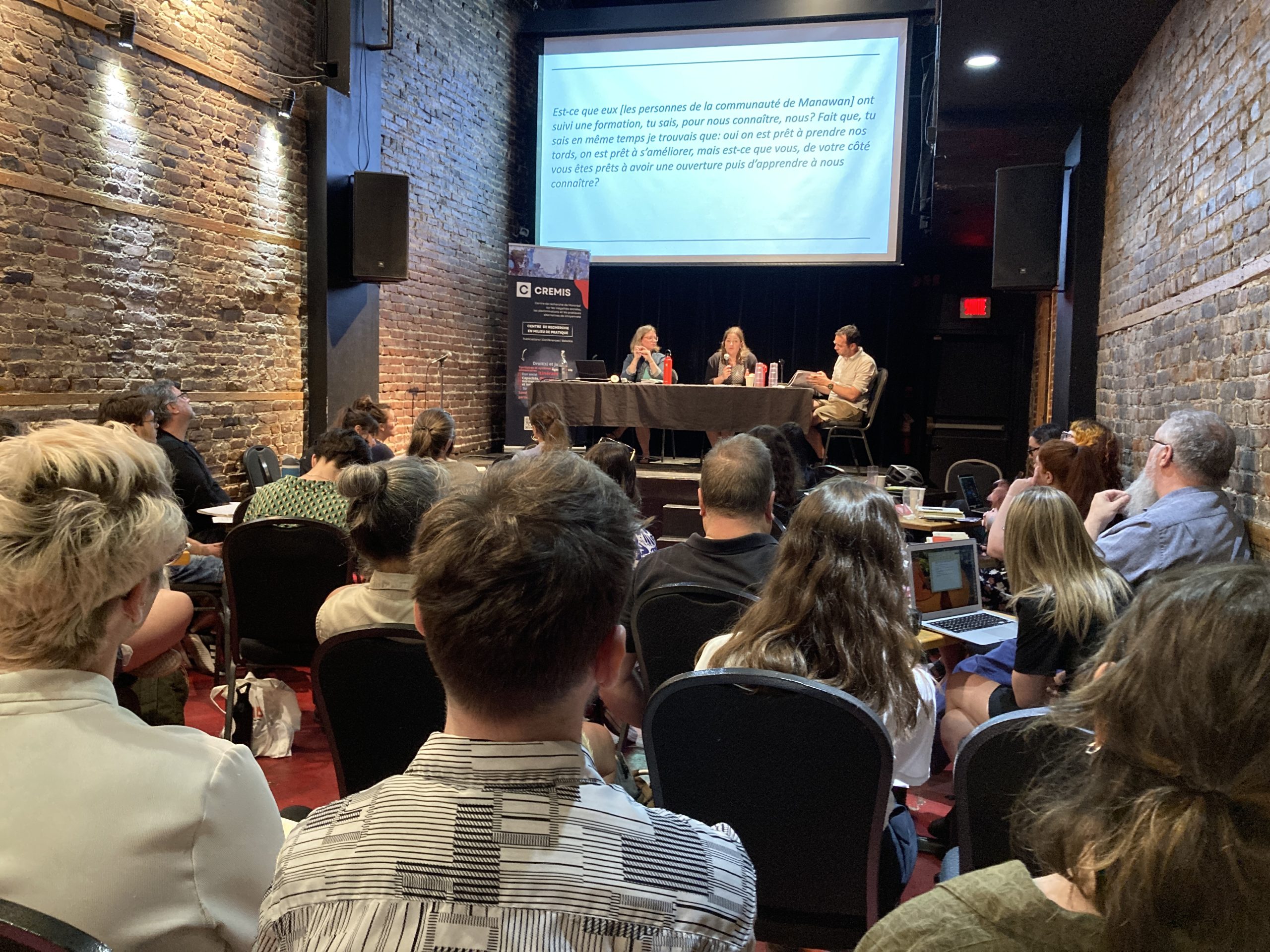 Au Théâtre Sainte-Catherine, lors de l'événement « Paternalisme, actualité et contestations ». Au premier plan, les participant·e·s à l'événement, de dos. à l'arrière plan, deux chercheuses présentent leur recherche, aux côté de l'organisateur de l'événement.