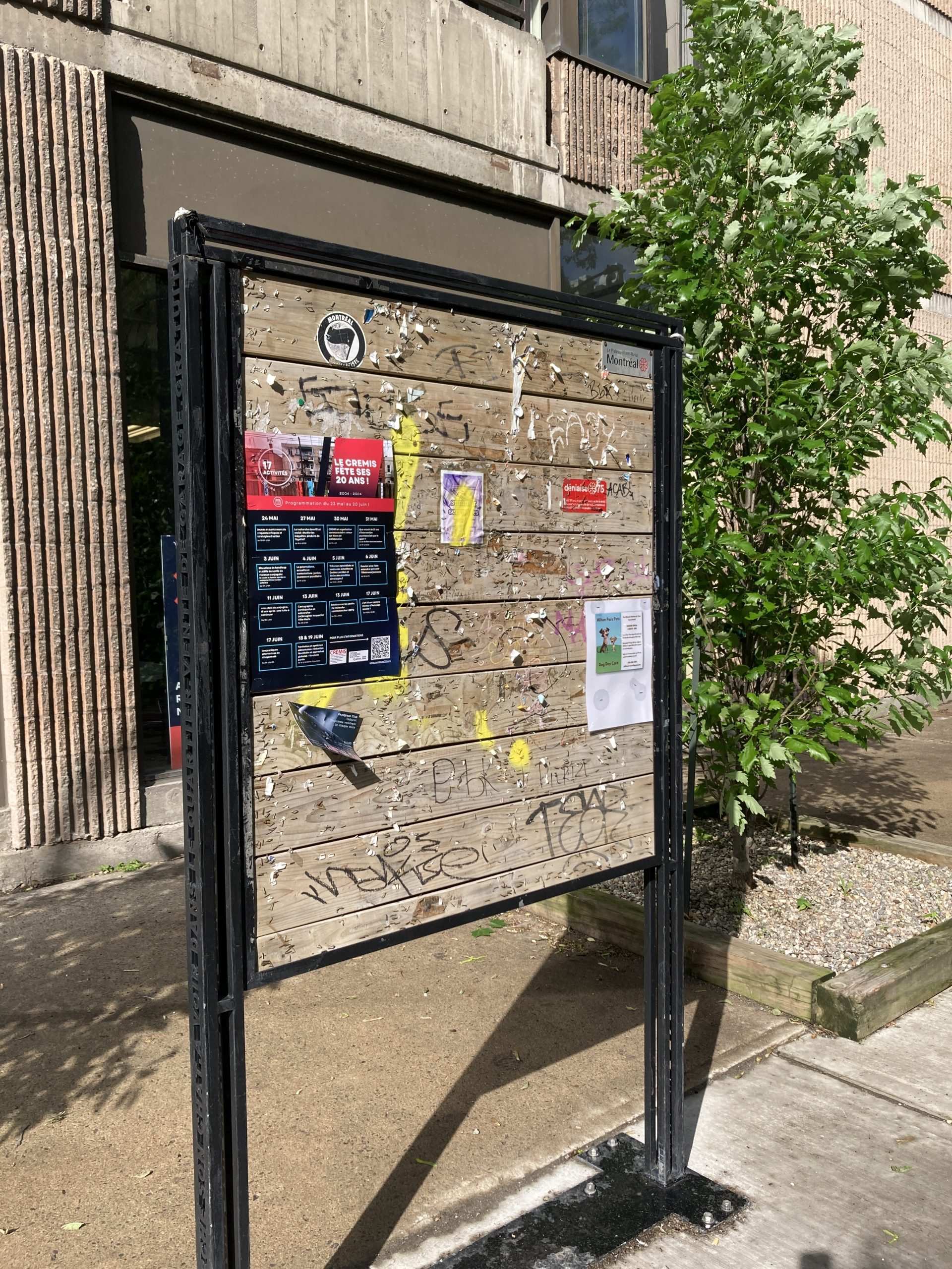 Affiche de la programmation des 20 ans du CREMIS sur un babillard dans la rue. Le babillard contient des restants d'affiches semi déchirée et des graffitis. 